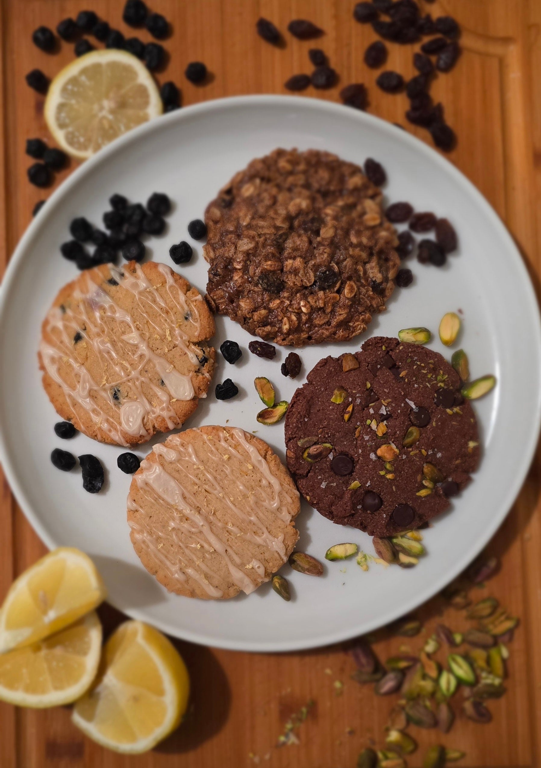 4 different cookies (lemon cookie with lemon glaze, lemon blueberry cookie with lemon glaze, double chocolate pistachio cookies with chocolate chips and chopped pistachios, oatmeal raisin cookie) on a plate with raisins and pistachios, surrounded by lemons on a wooden surface. 