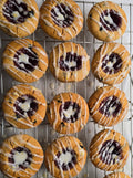 1 dozen lemon blueberry thumbprint cookies filled with red fruit preserves,  and freshly finished with a zesty lemon glaze on a cooling rack