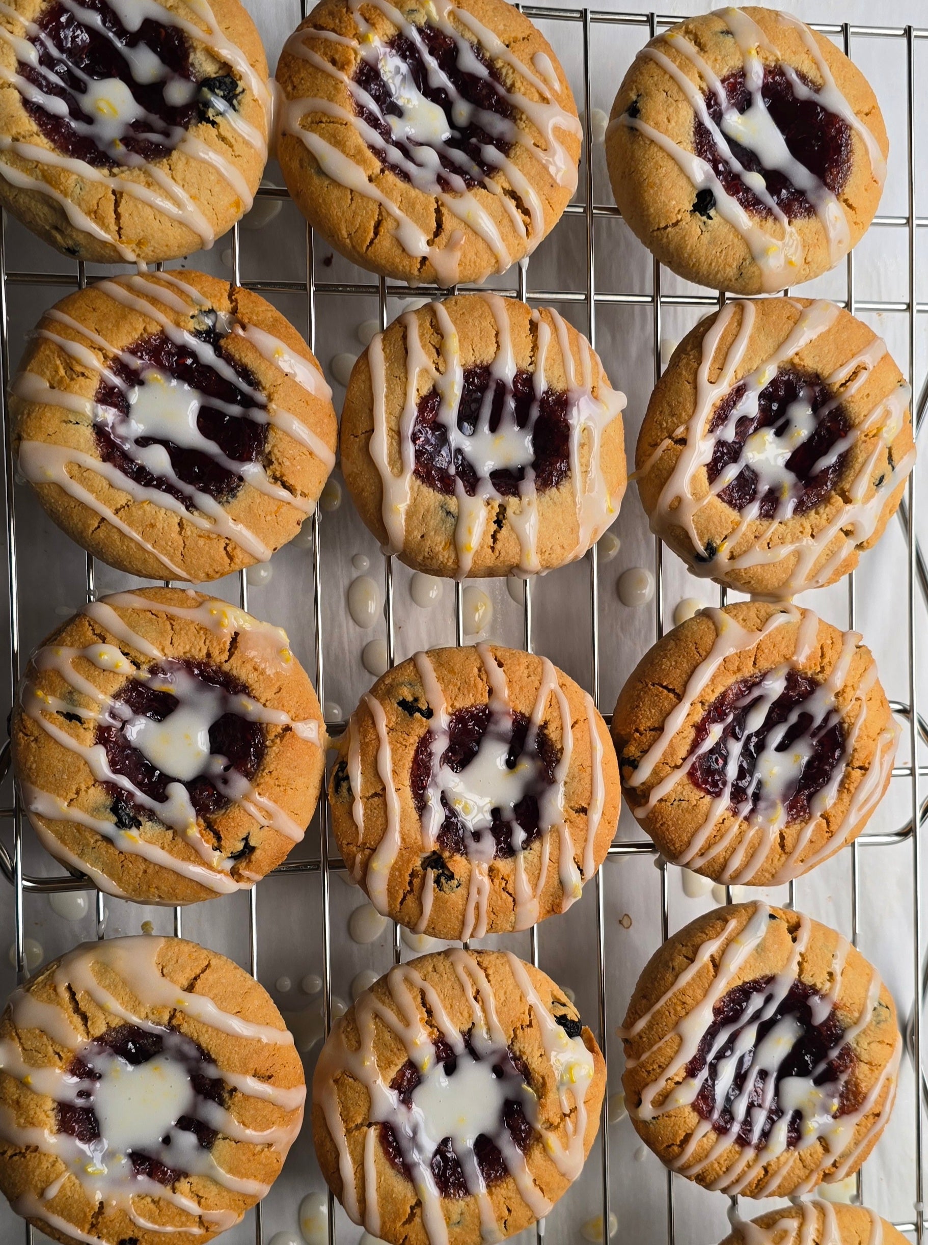 1 dozen lemon blueberry thumbprint cookies filled with red fruit preserves,  and freshly finished with a zesty lemon glaze on a cooling rack