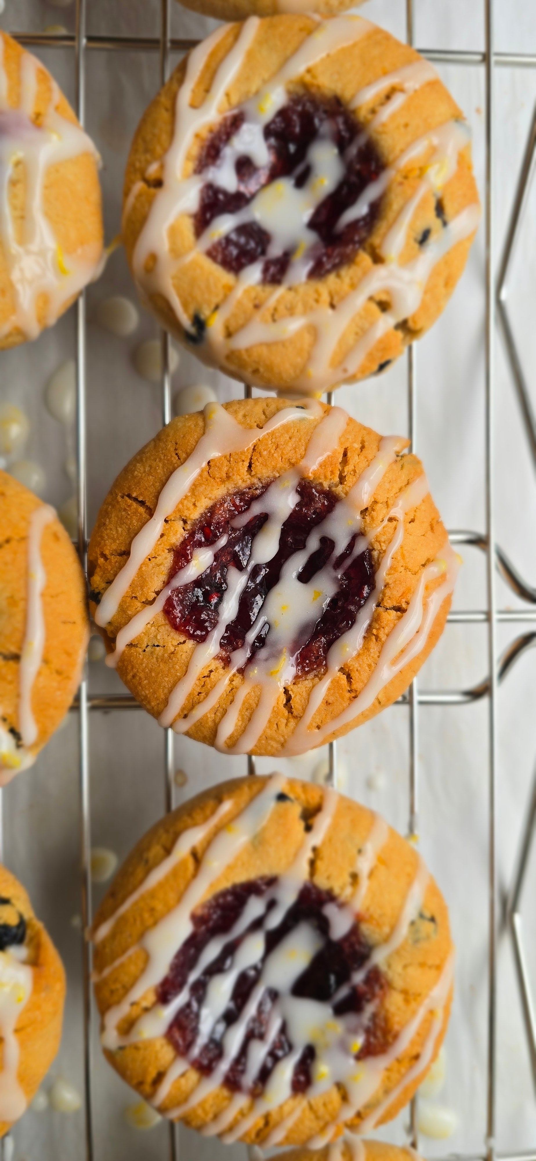 3 lemon blueberry thumbprint Cookies with white lemon drizzle and red fruit preserve filling on a cooling rack