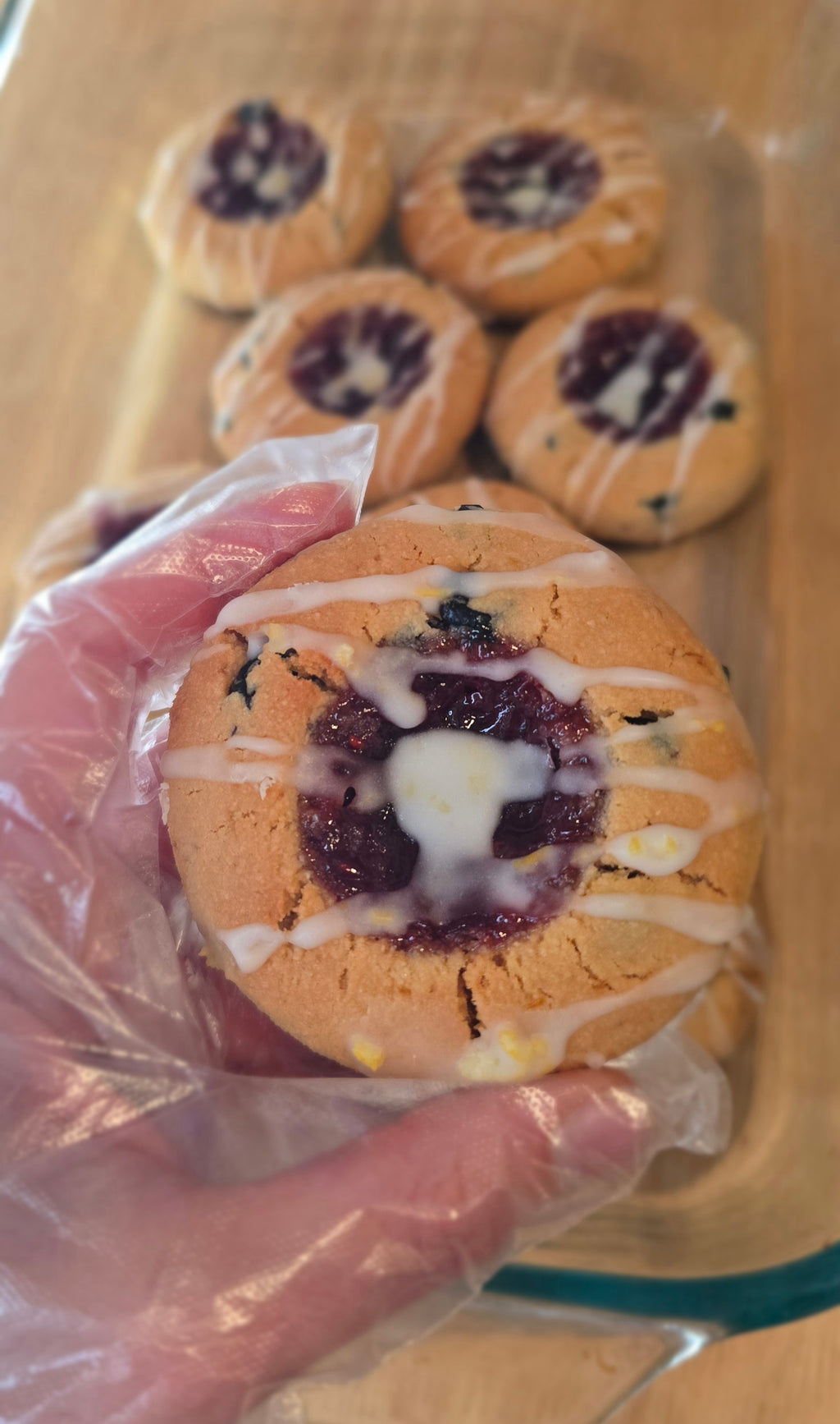 gloved hand holding a lemon blueberry thumbprint cookie with red fruit preserve filling and white lemon drizzle over a tray of similar cookies.