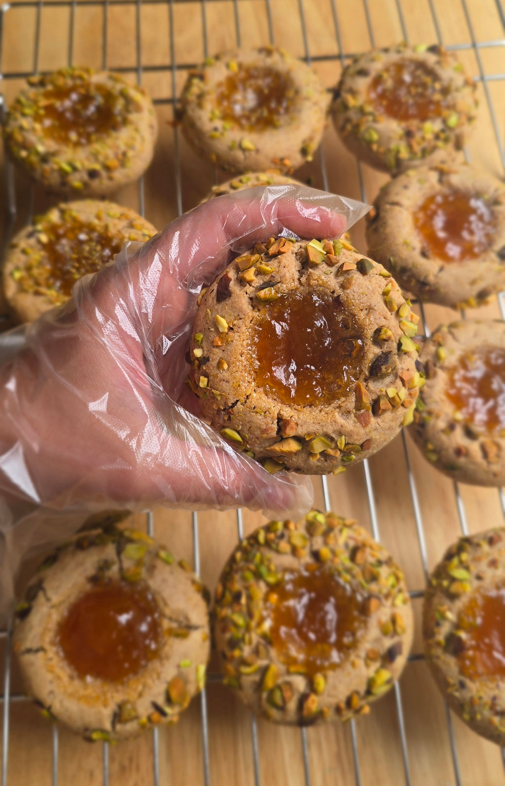 A hand wearing a clear plastic glove holds a pistachio-topped apricot thumbprint cookie with a glossy jam center, with more cookies cooling on a wire rack in the background.