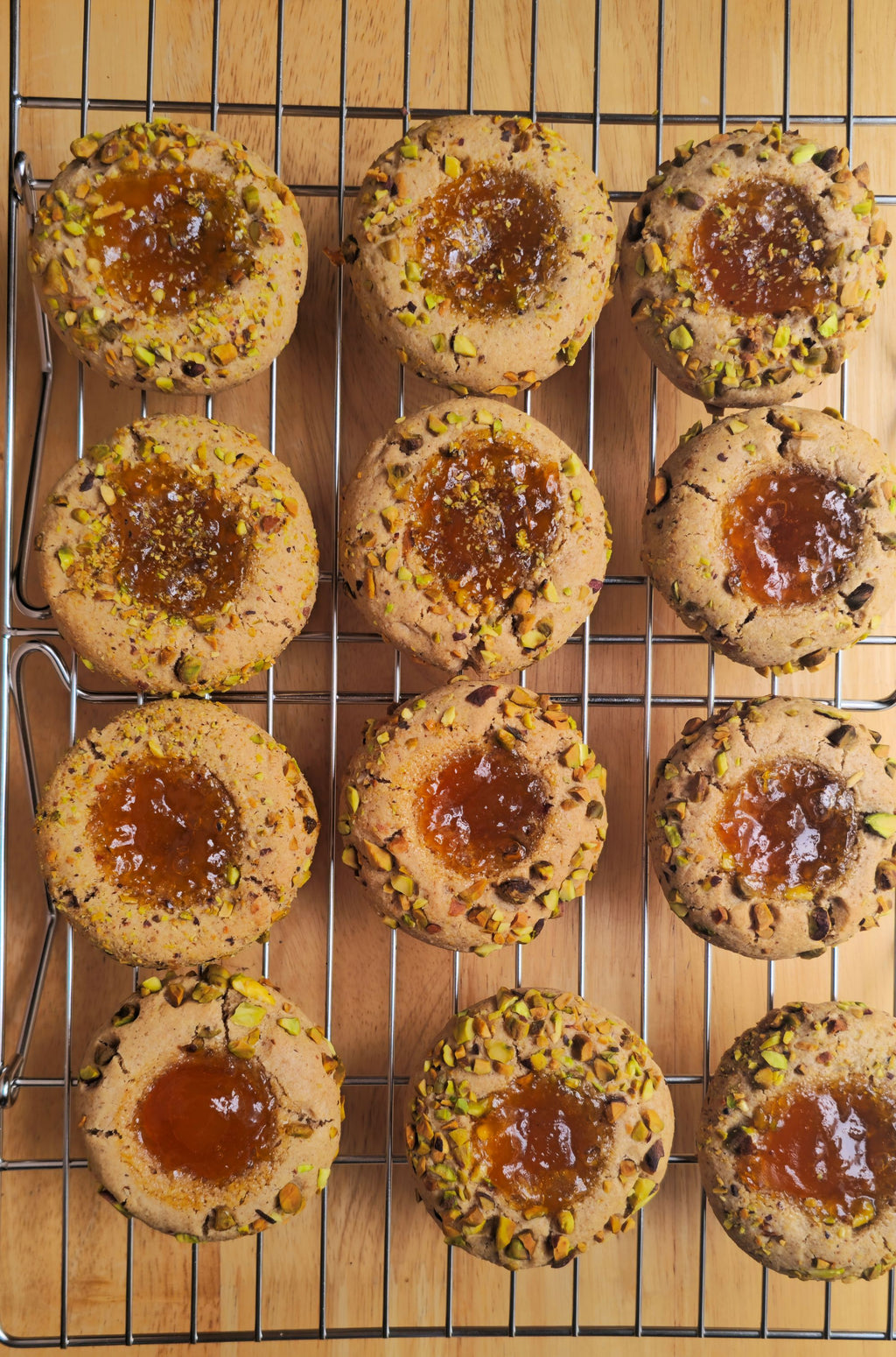 Twelve thumbprint cookies with apricot jam centers and chopped pistachio topping, arranged on a metal cooling rack over a wooden surface.