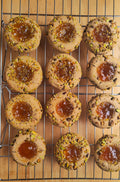 Twelve thumbprint cookies with apricot jam centers and chopped pistachio topping, arranged on a metal cooling rack over a wooden surface.