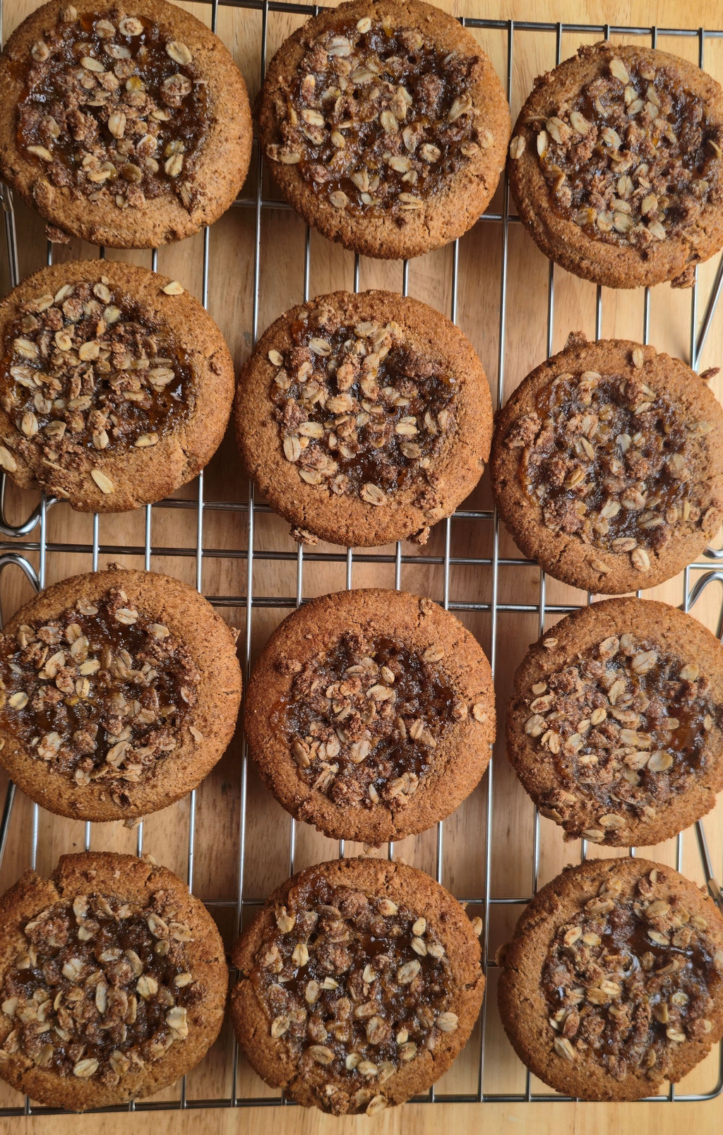 Twelve golden-brown cookies filled with peach preserves, and topped with rolled oats streusel on a metal cooling rack over a light wood surface.
