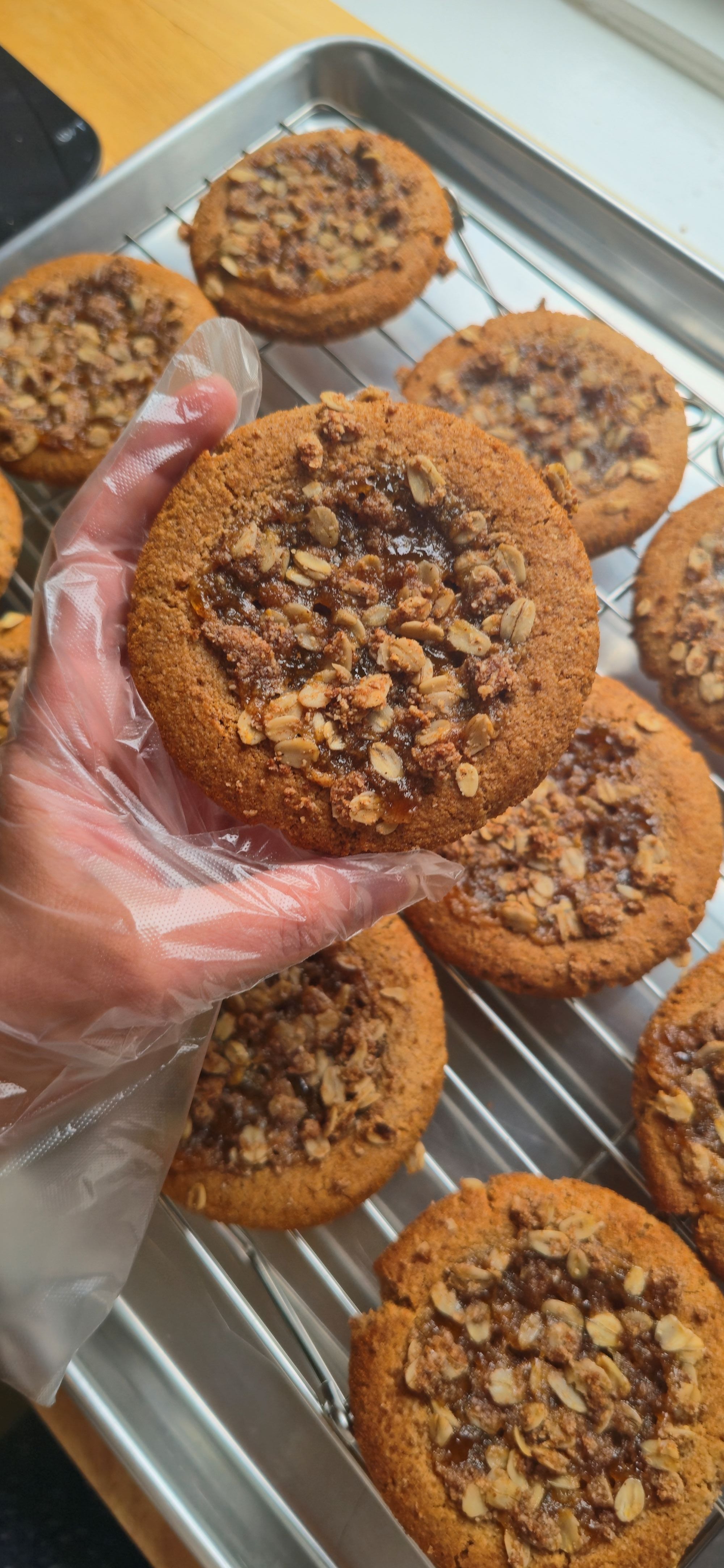 A hand wearing a clear plastic glove holds a freshly baked peach cobbler snickerdoodle cookie on a metal cooling rack, with more cookies visible in the background.