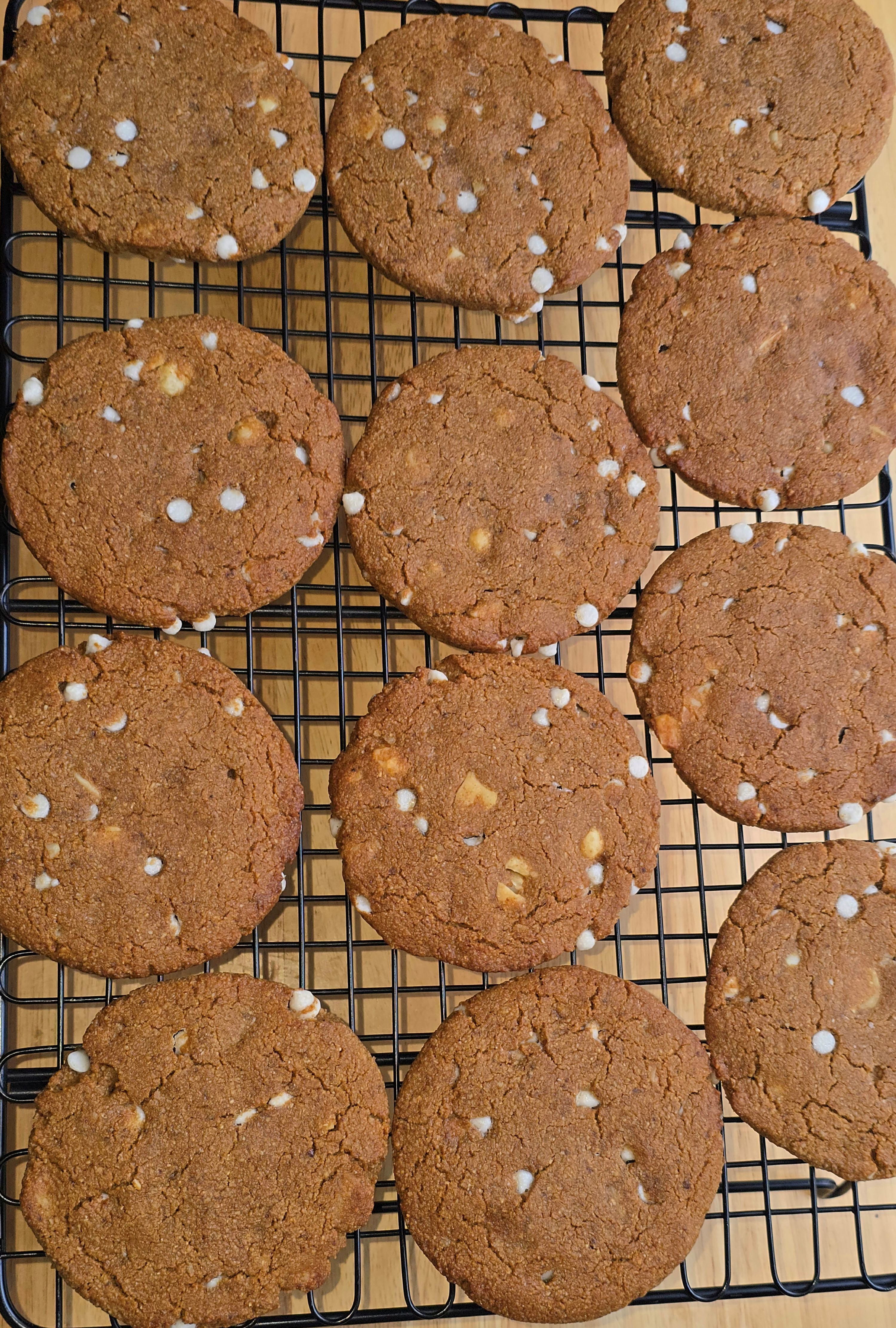 12 Cookies with white chocolate chips and macadamia nuts laying flat on a black cooling rack that is on a wooden surface. 